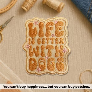 May include: Embroidered patch with the text "LIFE IS BETTER WITH DOGS" in a tan color with pink floral accents. The patch has a scalloped edge and a dog paw print. The patch is surrounded by sewing supplies.
