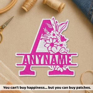 May include: A hot pink embroidered patch in the shape of the letter A, featuring white floral accents and a horizontal bar across the center with the text ANYNAME in hot pink. The patch has a white border and a textured, stitched fabric finish.