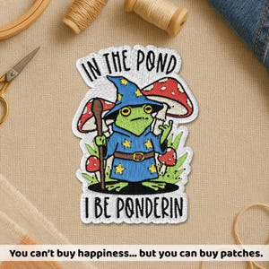 May include: Embroidered patch featuring a frog wizard with the text "IN THE POND" and "I BE PONDERIN". The frog wears a blue wizard hat and robe with gold stars. Red and white mushrooms and green foliage surround the frog.