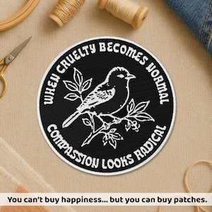 Peut inclure: Écusson brodé noir et blanc avec un oiseau sur une branche et le texte "When Cruelty Becomes Normal, Compassion Looks Radical". Le patch est circulaire avec une bordure blanche. Le texte "You can't buy happiness... but you can buy patches" est en bas.