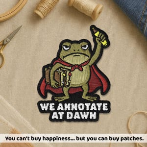 May include: An embroidered patch featuring a green frog wearing a red cape, holding a brown book and a yellow pencil. The text below the frog reads WE ANNOTATE AT DAWN in white capital letters on a black background with a thick black border.