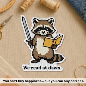 May include: Embroidered patch featuring a cartoon raccoon holding a sword and a book. The raccoon is brown, black, and white, with the text "We read at dawn." The patch is on a beige background.