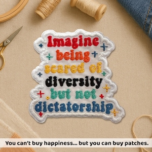 Peut inclure: Écusson brodé avec le texte "Imagine being scared of diversity but not dictatorship" en rouge, orange, noir et bleu. L'écusson a une bordure blanche et des accents décoratifs en forme de croix et d'étoiles. Des fournitures de couture entourent l'écusson.