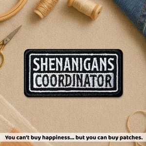 May include: A black and white embroidered patch with the text "SHENANIGANS COORDINATOR" in a rectangular shape. The patch is surrounded by a black border. The background includes sewing supplies and the text: "You can't buy happiness... but you can buy patches."
