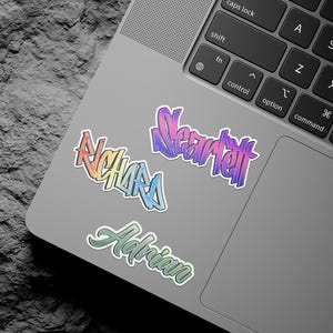 May include: A silver laptop computer with three colorful stickers on the lid. The stickers are in a gradient of pink, purple, blue, and green. The stickers say "Near tet", "Adrian", and "Richards".