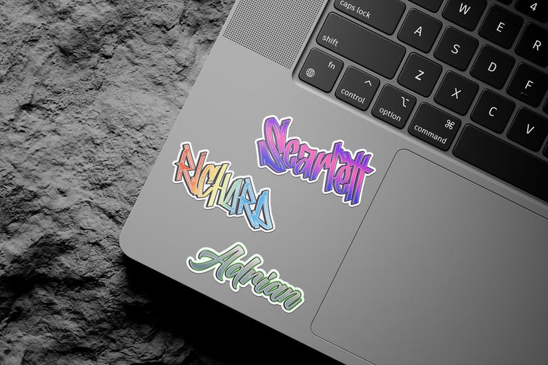 May include: A silver laptop computer with three colorful stickers on the lid. The stickers are in a gradient of pink, purple, blue, and green. The stickers say "Near tet", "Adrian", and "Richards".