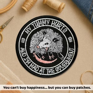May include: A round, black patch with white embroidered text that reads "MY TUMMY HURTS AND I'M MAD AT THE GOVERNMENT." The patch features a detailed opossum illustration. The text "You can't buy happiness... but you can buy patches" is at the bottom.