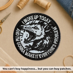 May include: A round, black and white embroidered patch featuring a snarling opossum. The text around the opossum reads "I WOKE UP TODAY AND I'M GONNA MAKE IT EVERYONE'S PROBLEM". The patch is surrounded by sewing supplies.