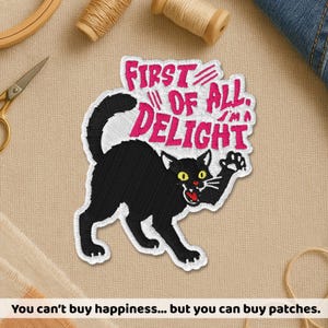 May include: Embroidered patch featuring a black cat with yellow eyes and an open mouth, arched back, and raised paw. The patch has the text "FIRST OF ALL, I'M A DELIGHT" in pink. The patch is on a beige background.