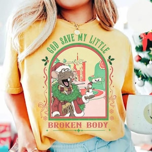 May include: Yellow t-shirt with a graphic featuring a cartoon rat in a Santa hat, holding a snowman. The graphic is framed with a red and green border and includes the text "GOD SAVE MY LITTLE BROKEN BODY".