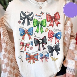May include: Cream-colored t-shirt featuring colorful bow designs inspired by Marvel characters like Spider-Man, Captain America, and Venom. Each bow is uniquely colored and styled, with character names above. A fun and creative graphic tee.