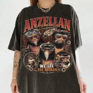 May include: A black short-sleeve t-shirt featuring a graphic print of three Anzellan characters with goggles. The text reads "ANZELLAN Droidsmiths" and "WE FIX THE BROKEN" with small circular emblems. The shirt has a faded, washed-out appearance.
