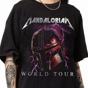 May include: Black t-shirt featuring a graphic of a Mandalorian helmet with purple lightning and the text "MANDALORIAN" and "THIS IS THE WAY". Below the helmet is the text "WORLD TOUR" in white.