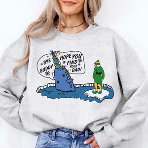 Puede incluir: Una sudadera gris con un gráfico navideño que presenta un narval con una cadena de luces y el texto "Bye Buddy" y un elfo con el texto "¡Espero que encuentres a tu papá!"