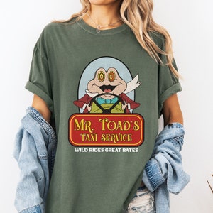 Könnte beinhalten: Grünes T-Shirt mit einer Vintage-Cartoon-Illustration eines Frosches, der ein Auto fährt. Der Text auf dem Shirt lautet "Mr. Toad's Taxi Service. Wild Rides Great Rates."