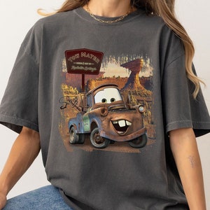 Może przedstawiać: Szary T-shirt z grafiką Tow Matera z filmu Disneya Pixara Auta. Grafika przedstawia Tow Matera jadącego drogą ze znakiem, na kt&oacute;rym widnieje napis "Tow Mater - World's Best Tow Truck - Radiator Springs".