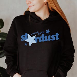 Könnte beinhalten: Schwarzer Kapuzenpullover mit einer Vordertasche und dem Wort "Stardust" in blauer Schrift mit einer hellblauen Sternengrafik. Der Text "Freya Skye" steht über dem Wort "Stardust".