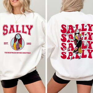 K&ouml;nnte beinhalten: Wei&szlig;er Pullover mit einem roten und schwarzen Aufdruck von Sally aus Nightmare Before Christmas. Die Vorderseite des Pullovers tr&auml;gt die Aufschrift "Sally Est. 1993 The Nightmare Before Christmas". Die R&uuml;ckseite des Pullovers zeigt das Wort "Sally" viermal in einer roten Retro-Schrift.