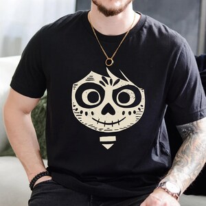 Könnte beinhalten: Schwarzes T-Shirt mit einem weißen Aufdruck eines Cartoon-Skelett-Charakters mit einem breiten Grinsen. Der Charakter hat ein Schädelgesicht mit großen Augen und einem kleinen Mund. Der Charakter trägt einen Hut.