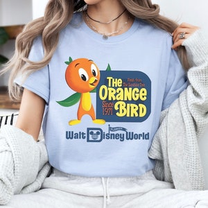 Puede incluir: Una camiseta azul claro con un pájaro naranja de dibujos animados que lleva una hoja verde. El texto de la camiseta dice "The Orange Bird" con las palabras "Fresh from the Sunshine Tree" y "Since 1971" en letra más pequeña. El texto "Walt Disney World" está debajo del pájaro con la palabra "Florida" en letra más pequeña.