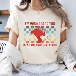 May include: A white t-shirt with a retro-style graphic of a cartoon devil holding a pitchfork. The text "I'm gonna lead you down the path that rocks" is printed on the shirt, along with the signature "K.B."