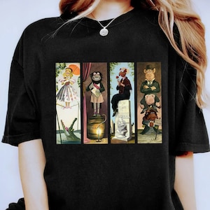 Puede incluir: Camiseta negra con un estampado gráfico de cuatro personajes de los Muppets de la película The Great Muppet Caper. Los personajes son Miss Piggy, Animal, Sam el Águila y Scooter. Los personajes están de pie en diferentes plataformas, con el texto "Dinamita", "Descansa en paz, queridos amigos, nunca te olvidaremos, George", y "Arena movediza" visible.