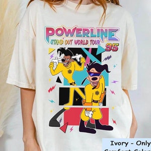 May include: A white t-shirt featuring a colourful graphic of two cartoon characters in yellow and black costumes. The text "POWERLINE STAND OUT WORLD TOUR 95" is printed on the shirt.