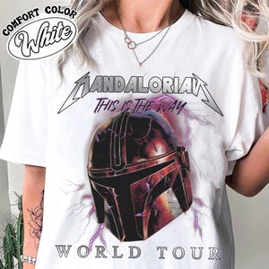 May include: White t-shirt featuring a graphic of a Mandalorian helmet with the text "Mandalorian This Is The Way World Tour". The design includes purple lightning bolts and a distressed effect.