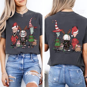 K&ouml;nnte beinhalten: Dunkelgraues T-Shirt mit einem festlichen Cartoon-Design. Auf der Vorder- und R&uuml;ckseite des Shirts sind drei Charaktere in festlicher Kleidung zu sehen, darunter Weihnachtsm&uuml;tzen und Rentiergeweihe. Das Design umfasst verpackte Geschenke und Lichterketten.