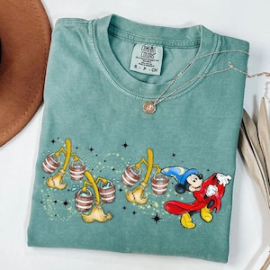 May include: A teal Comfort Colors t-shirt with a graphic of Mickey Mouse as a sorcerer, with animated brooms carrying buckets of water. The shirt has a crew neck and short sleeves. A silver necklace with a pendant is also visible.