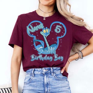 Puede incluir: Una camiseta granate con un personaje de serpiente de dibujos animados en un dise&ntilde;o azul con forma de Mickey Mouse. El texto incluye "Name", "5", "Smith" y "Birthday Boy" en azul y blanco. La camiseta la lleva una persona.