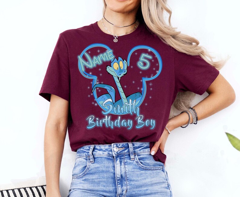 Puede incluir: Una camiseta granate con un personaje de serpiente de dibujos animados en un dise&ntilde;o azul con forma de Mickey Mouse. El texto incluye "Name", "5", "Smith" y "Birthday Boy" en azul y blanco. La camiseta la lleva una persona.