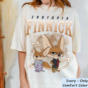 May include: Ivory comfort colour t-shirt with the word "Zootopia" above "Finnick" in gold. The shirt features a cartoon image of two foxes. The text "Ivory - Only Comfort Color" is at the bottom of the shirt.