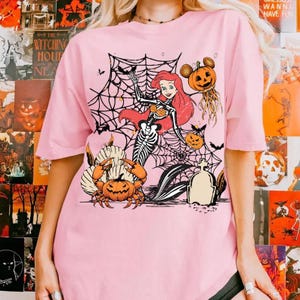 May include: Pink t-shirt featuring a Halloween-themed graphic. The design includes a skeleton mermaid, spiderwebs, pumpkins, bats, and a Mickey Mouse-inspired pumpkin. The shirt has short sleeves and a crew neck.