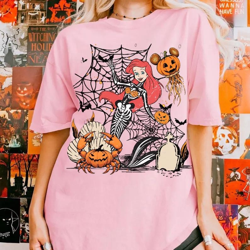 May include: Pink t-shirt featuring a Halloween-themed graphic. The design includes a skeleton mermaid, spiderwebs, pumpkins, bats, and a Mickey Mouse-inspired pumpkin. The shirt has short sleeves and a crew neck.