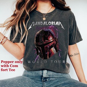 May include: Dark gray t-shirt featuring a graphic of a Mandalorian helmet with purple lightning and the text "MANDALORIAN THIS IS THE WAY WORLD TOUR". The shirt is a comfort tee.
