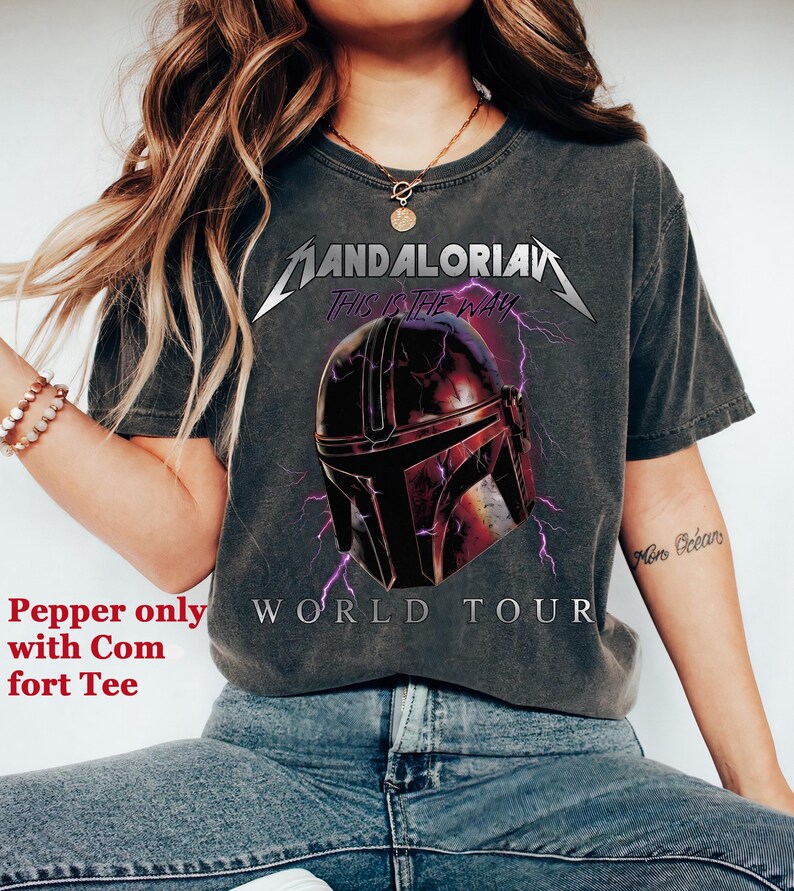 May include: Dark gray t-shirt featuring a graphic of a Mandalorian helmet with purple lightning and the text "MANDALORIAN THIS IS THE WAY WORLD TOUR". The shirt is a comfort tee.
