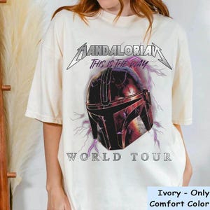 May include: Ivory-colored t-shirt featuring a graphic of a Mandalorian helmet with lightning bolts and the text "Mandalorian World Tour." The phrase "This is the Way" is above the helmet. The shirt is a Comfort Color brand.