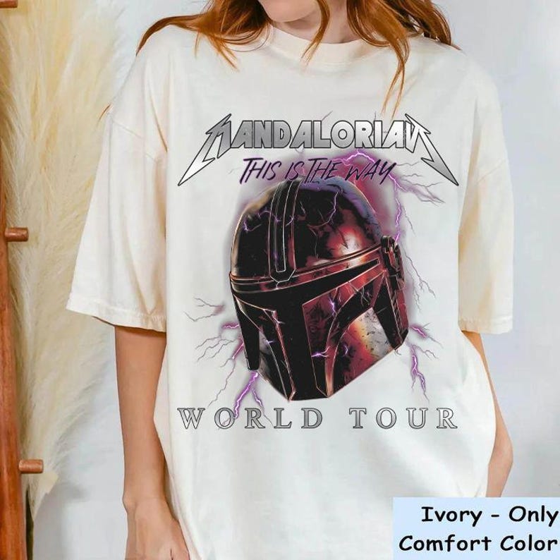 May include: Ivory-colored t-shirt featuring a graphic of a Mandalorian helmet with lightning bolts and the text "Mandalorian World Tour." The phrase "This is the Way" is above the helmet. The shirt is a Comfort Color brand.