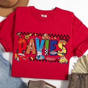 May include: A red t-shirt featuring the name "DAVIES" in colorful, cartoon-style lettering with car-themed illustrations. The design includes images of various animated cars and Route 66 signage.