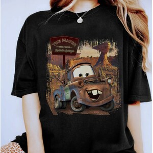 Może przedstawiać: Czarny T-shirt z grafiką Tow Matera z filmu Disneya Pixara Auta. Grafika przedstawia Tow Matera jadącego po drodze gruntowej ze znakiem, na kt&oacute;rym widnieje napis "Tow Mater - Witamy w Radiator Springs".