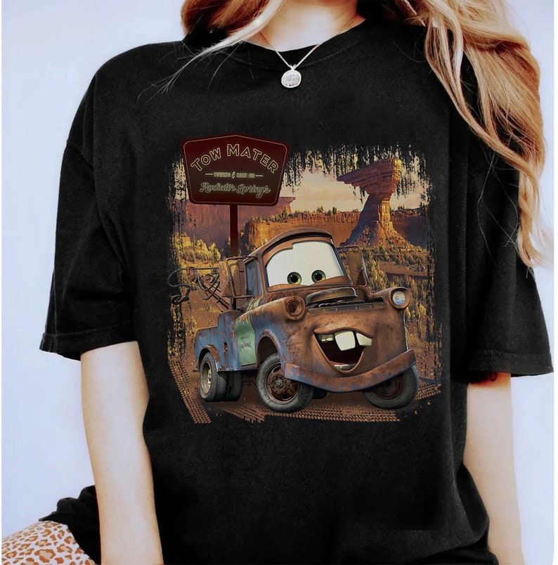 Może przedstawiać: Czarny T-shirt z grafiką Tow Matera z filmu Disneya Pixara Auta. Grafika przedstawia Tow Matera jadącego po drodze gruntowej ze znakiem, na kt&oacute;rym widnieje napis "Tow Mater - Witamy w Radiator Springs".
