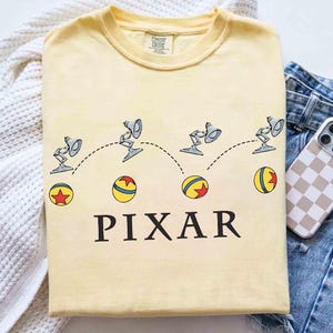 Pode incluir: Camiseta amarelo claro com um design Pixar. O gráfico mostra a lâmpada Pixar saltando sobre uma bola, com a palavra "PIXAR" impressa abaixo em preto. Um telefone em uma capa xadrez e jeans estão ao lado da camiseta.