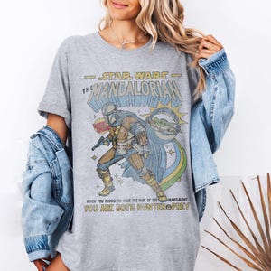K&ouml;nnte beinhalten: Ein graues T-Shirt mit einer Grafik im Stil eines Vintage-Comics eines Mandalorianer-Charakters aus Star Wars. Der Text auf dem Shirt lautet "Star Wars - The Mandalorian" und "Wenn du dich entscheidest, den Weg des Mandalorianers zu gehen, bist du sowohl J&auml;ger als auch Beute."