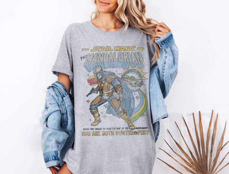 K&ouml;nnte beinhalten: Ein graues T-Shirt mit einer Grafik im Stil eines Vintage-Comics eines Mandalorianer-Charakters aus Star Wars. Der Text auf dem Shirt lautet "Star Wars - The Mandalorian" und "Wenn du dich entscheidest, den Weg des Mandalorianers zu gehen, bist du sowohl J&auml;ger als auch Beute."