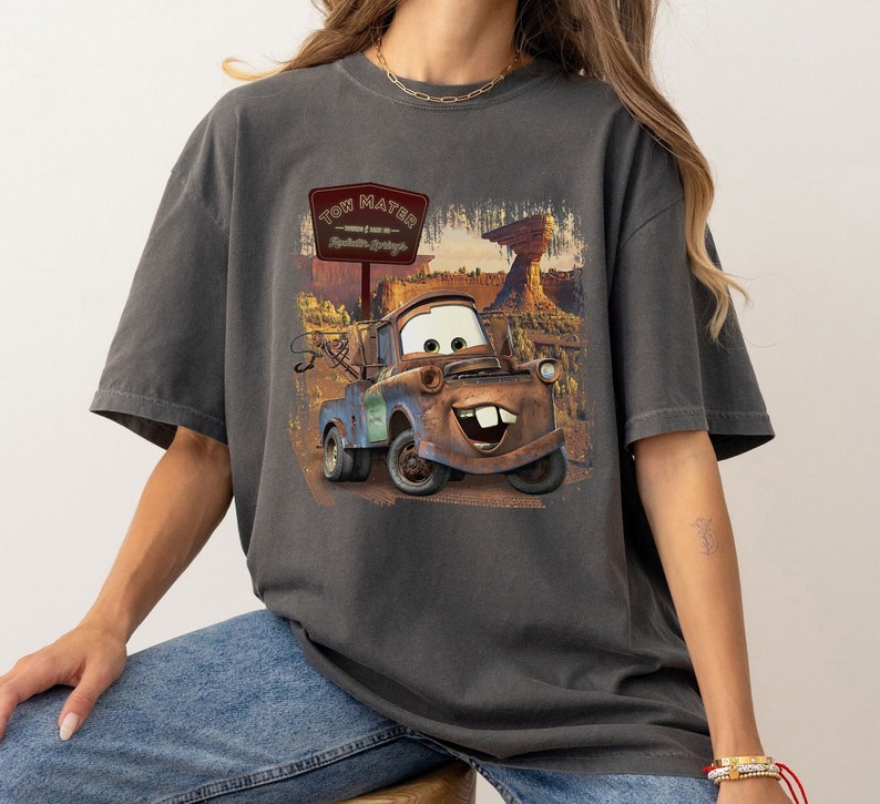 Może przedstawiać: Szary T-shirt z grafiką Tow Matera z filmu Disneya Pixara Auta. Grafika przedstawia Tow Matera jadącego drogą ze znakiem, na kt&oacute;rym widnieje napis "Tow Mater - World's Best Tow Truck - Radiator Springs".