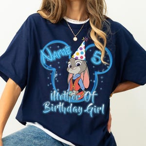 Puede incluir: Camiseta azul marino con una imagen de dibujos animados de Judy Hopps de Zootopia con un gorro de fiesta. La camiseta tiene el texto "Name 5 Mother of Birthday Girl" en fuente azul claro, con un contorno en forma de Mickey Mouse.