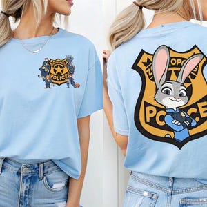 Puede incluir: Camiseta azul claro con un dise&ntilde;o de insignia de polic&iacute;a de dibujos animados en la espalda, con un oficial de conejo y el texto "Polic&iacute;a Zootr&oacute;polis". La parte delantera de la camiseta tiene un gr&aacute;fico de insignia de polic&iacute;a m&aacute;s peque&ntilde;o.