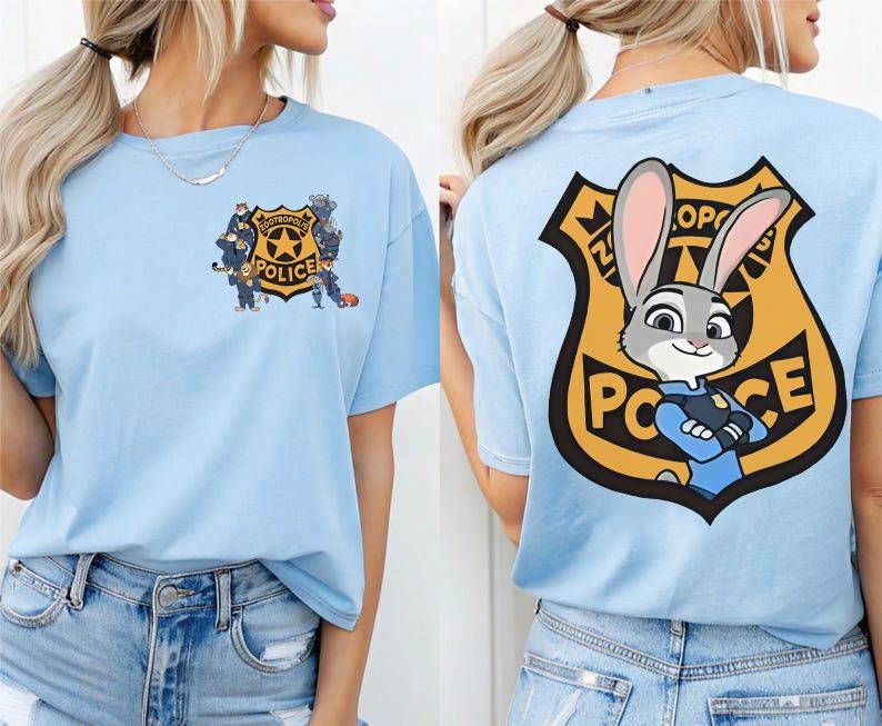 Puede incluir: Camiseta azul claro con un dise&ntilde;o de insignia de polic&iacute;a de dibujos animados en la espalda, con un oficial de conejo y el texto "Polic&iacute;a Zootr&oacute;polis". La parte delantera de la camiseta tiene un gr&aacute;fico de insignia de polic&iacute;a m&aacute;s peque&ntilde;o.