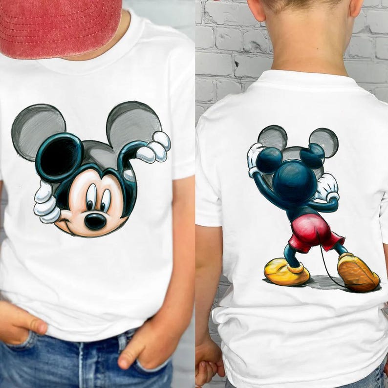 May include: White t-shirts featuring a Mickey Mouse graphic. One shirt shows Mickey's face, the other shows Mickey in a pose. The cartoon character is in black, blue, red, yellow, and white. The shirts are worn by a person.